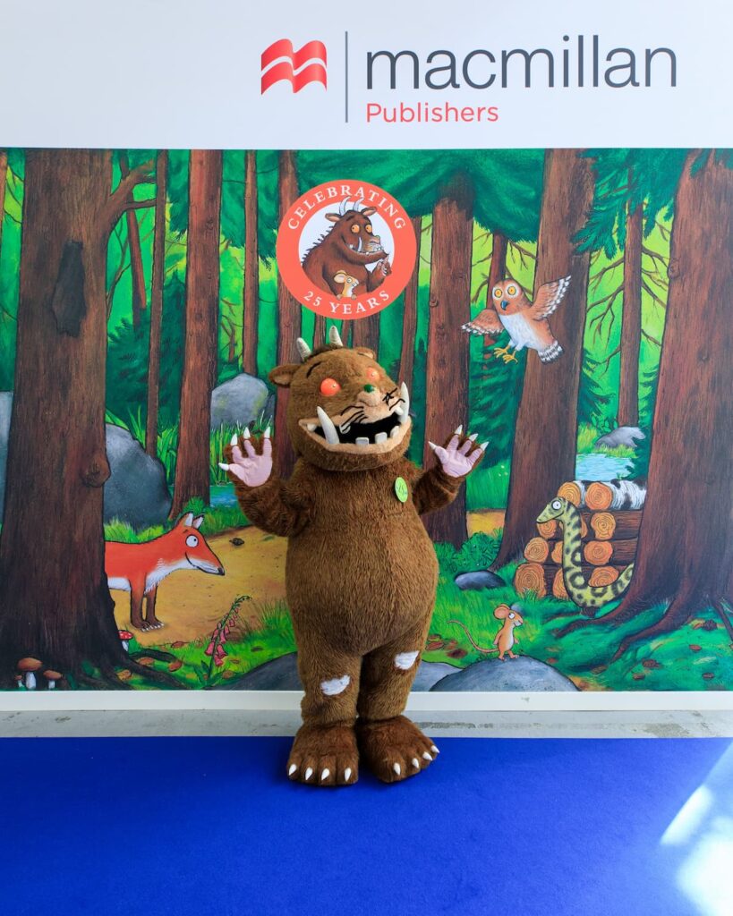 The Gruffalo character poses in front of the Macmillan Publishers Stand at Bologna Children's Book Fair, which features drawings from the original book by Axel Scheffler. The illustrations show a forest with a fox, owl, snake and mouse.