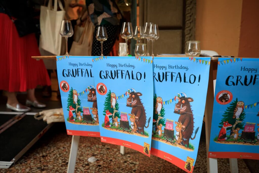 A close-up of branded The Gruffalo 25th anniversary posters stuck to the edge of a table holding glasses of wine.