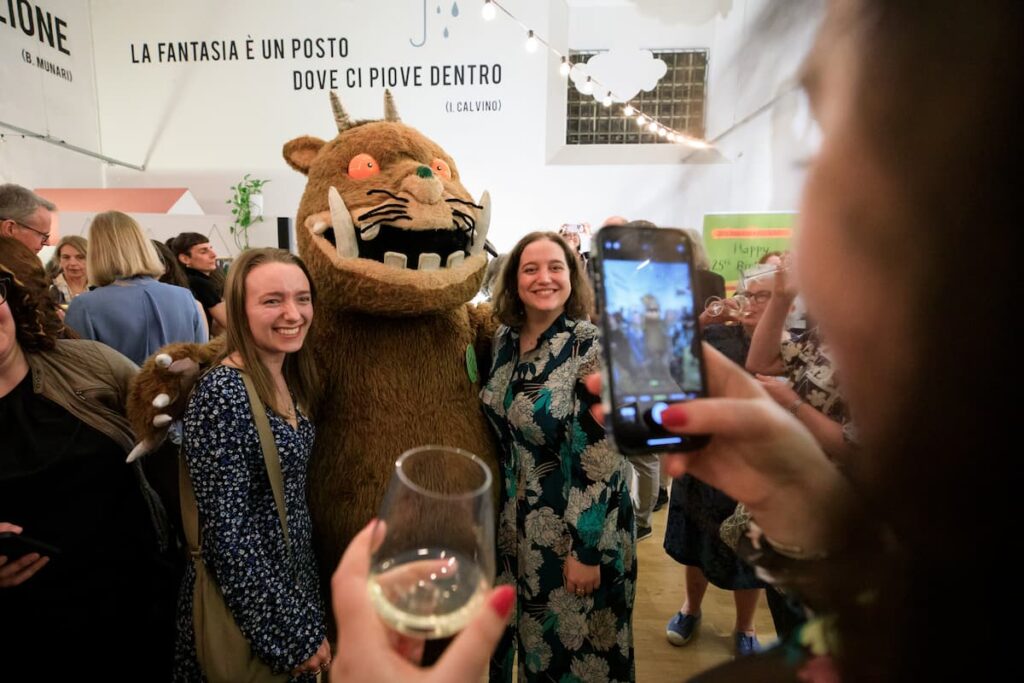 Two people pose with The Gruffalo character whilst another person takes a photograph in the foreground on a smartphone.