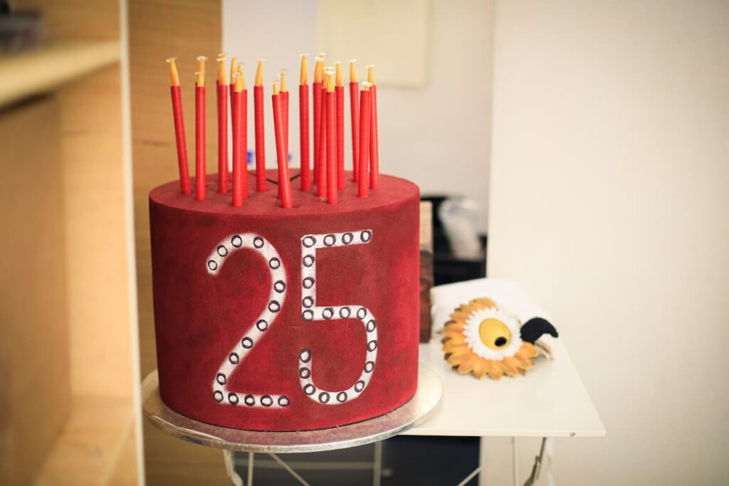 A bright red cake with '25' written on the front in white writing. The top is covered with 25 long red candles.