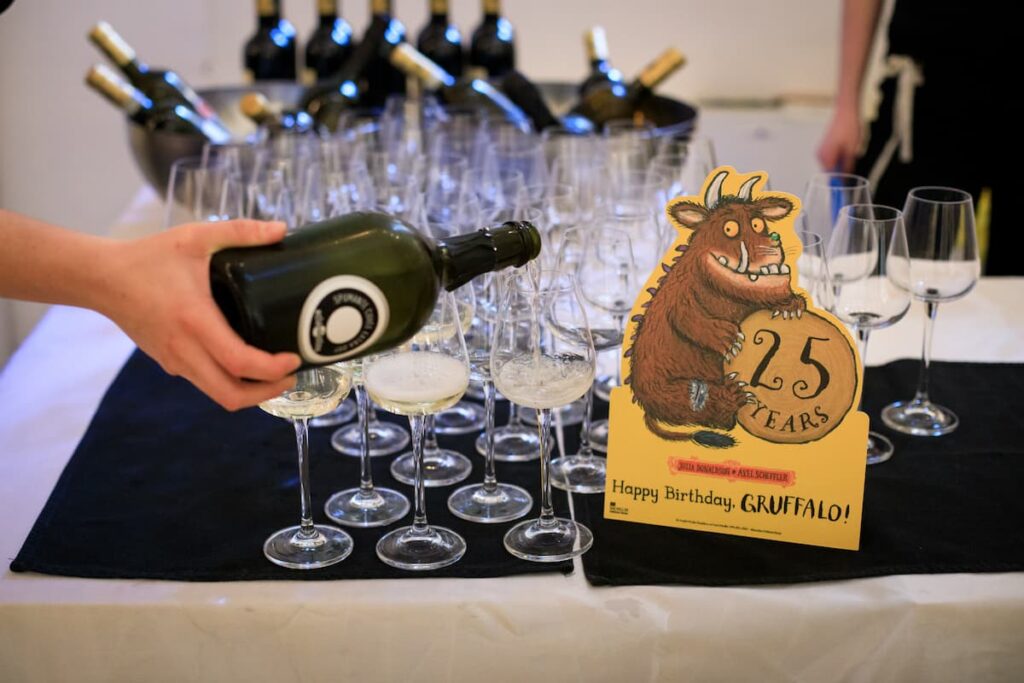A close-up of a glass of wine being poured next to a yellow standee of The Gruffalo holding a log with '25 years' written in the middle.