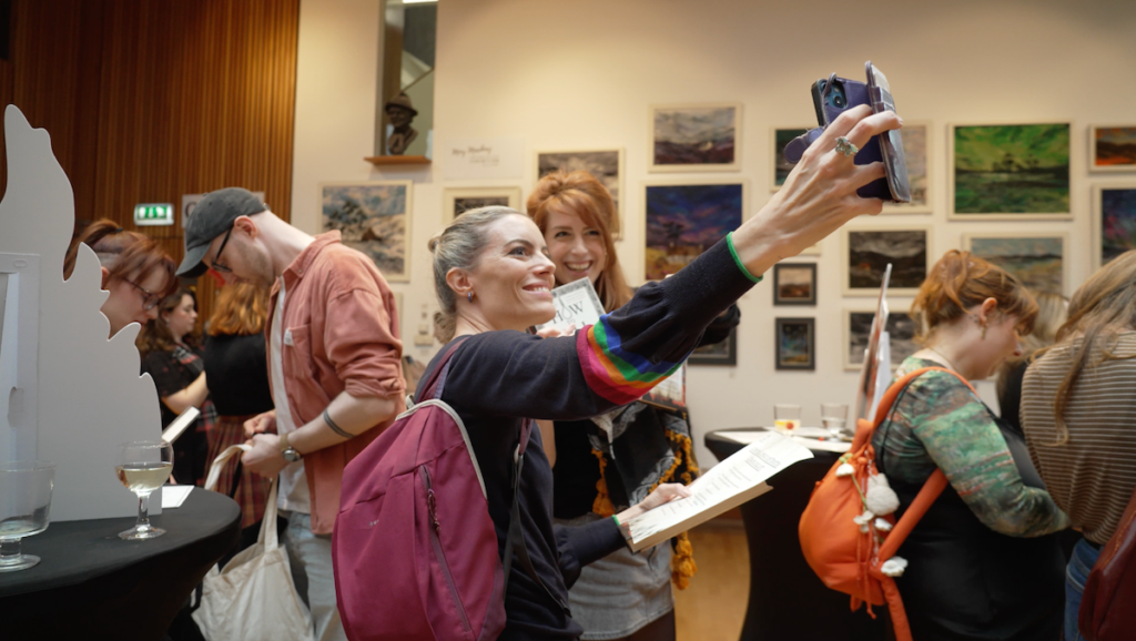 A woman with a purple backpack and rainbow-striped sleeve cuffs smiles while taking a phone selfie with a fellow influencers inside the event venue, which is lined with framed landscape paintings.