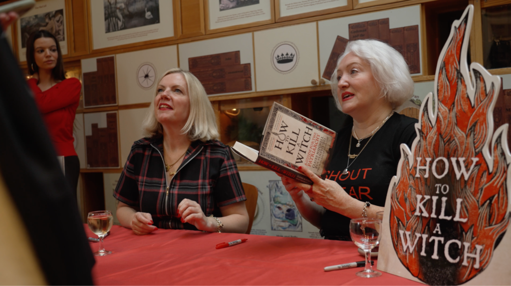 Authors Claire Mitchell and Zoe Venditozzi sitting side-by-side at a red tablecloth-draped table, signing copies of their book, How to Kill a Witch, with a flame-shaped promotional standee bearing the book title on the right.